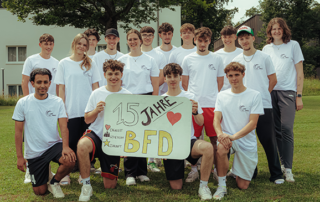 Die Freiwilligen stehen in weißen T-Shirts zum Gruppenfoto zusammen. Bei halten ein Schild mit "15 jahre BFD"